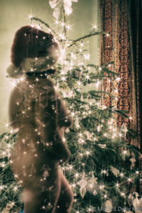 ghostly figure of nude woman in Santa hat in front of Christmas tree, with lights sparkling through her body