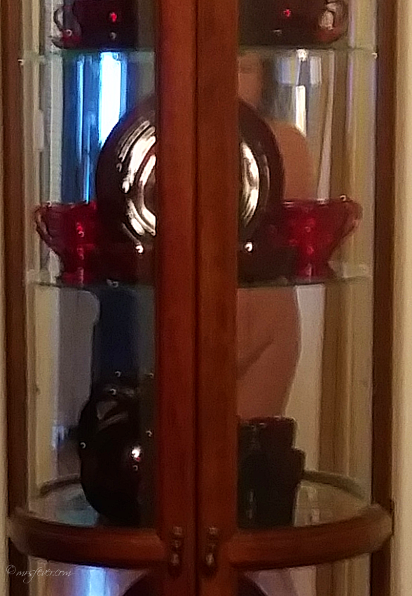woman's nude reflection caught in mirrored back of ruby red depression glass display case